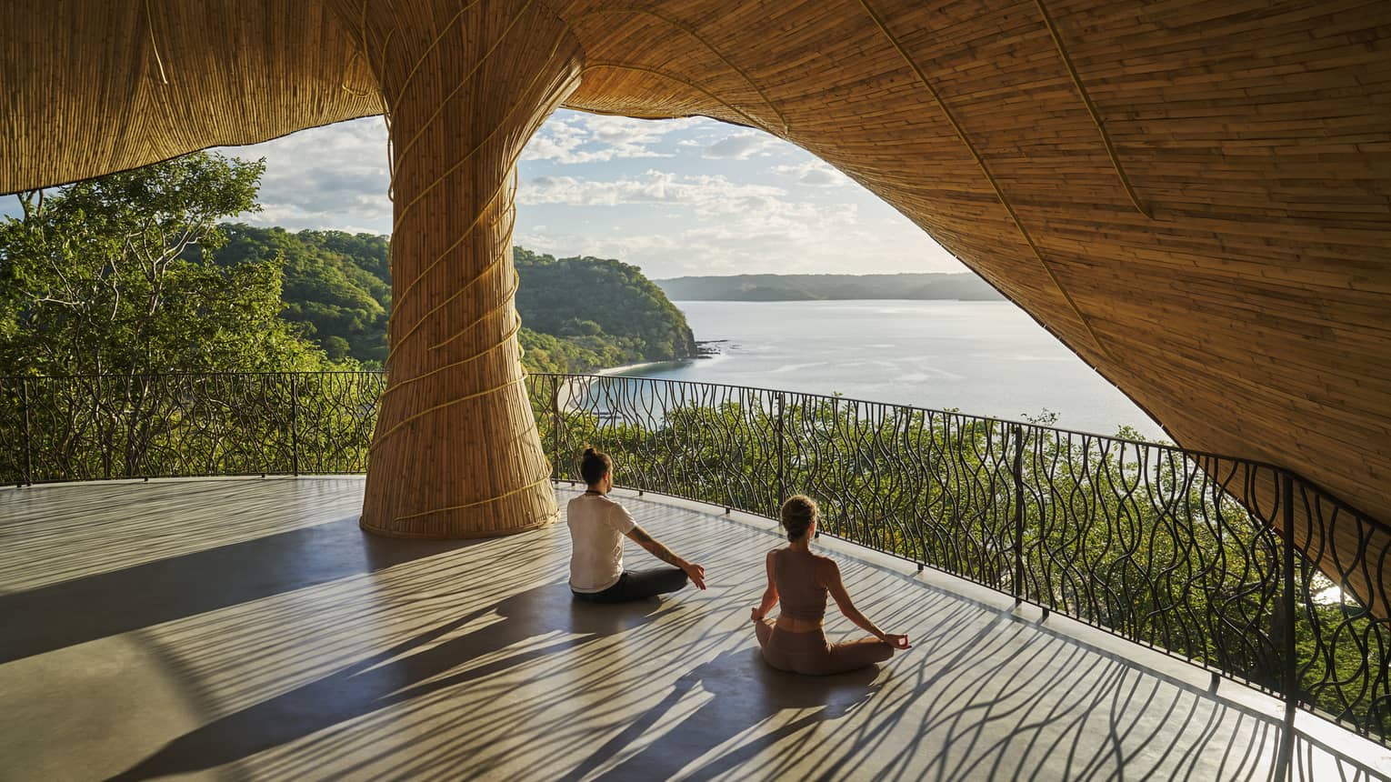 Two people sit with legs crossed and hands on knees on the floor of an open-air wellness studio that overlooks the ocean