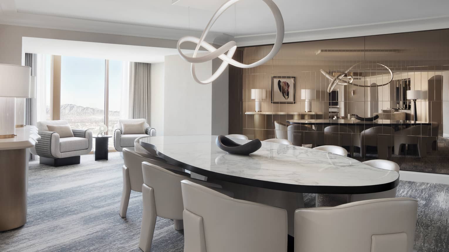 A modern dining room with white upholstered dining chairs and rounded marble table, an abstract chandelier and seating area behind by a large window overlooking a mountain view