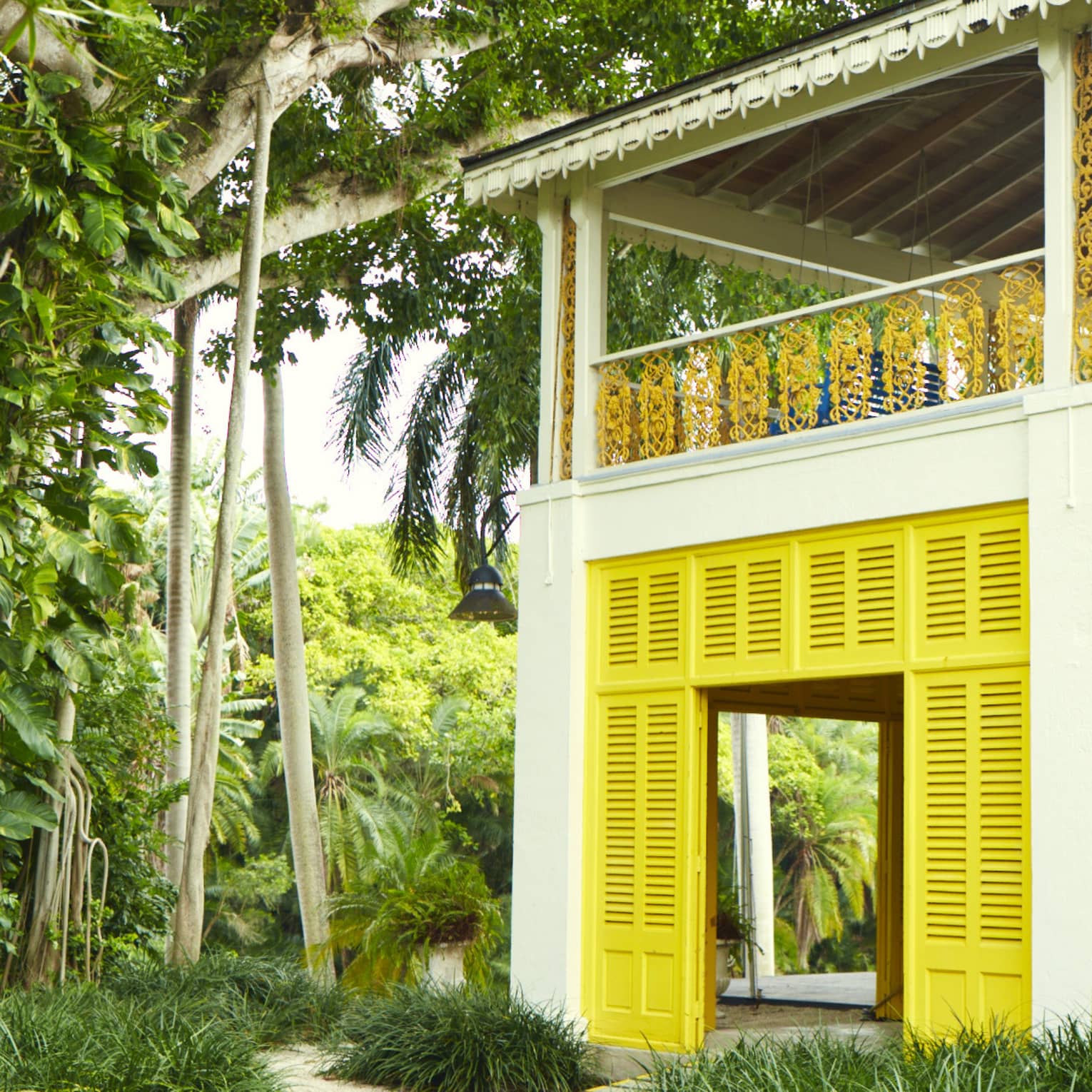 A cabana house with yellow shutter doors and a covered upper balcony with filigree railings is nestled amid lush foliage.
