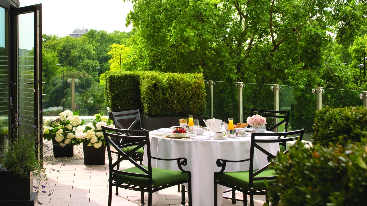 The Garden Suite at Four Seasons London Park Lane in Mayfair features a tranquil outdoor dining area overlooking lush parkland. The setting includes a well-appointed table dressed in white linen and set for breakfast with elegant china, fresh juice, and a variety of breakfast items. Comfortable green chairs surround the table, and the space is decorated with large potted plants and white hydrangeas, adding to the serene garden vibe. Glass railings ensure an unobstructed view of the green trees and foliage beyond, creating a peaceful and picturesque dining experience.
