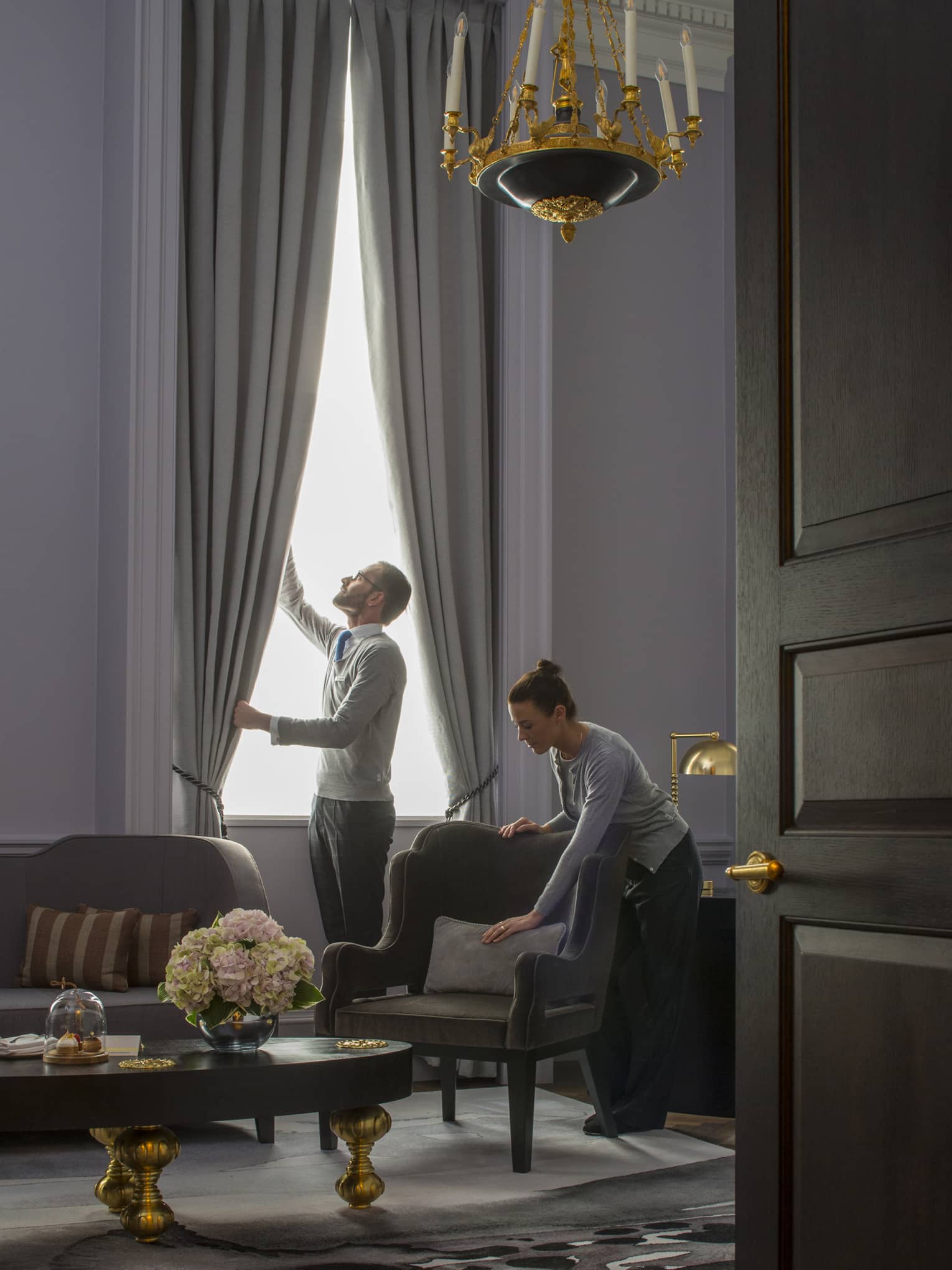 Grand Heritage Suite, man opens drapes as woman smooths pillow on armchair under chandelier