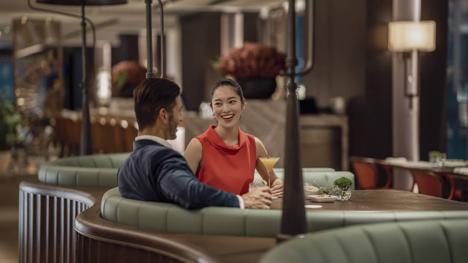A smiling couple sit at a circular booth in an empty, dimly lit restaurant with cocktails and appetizers on their table.