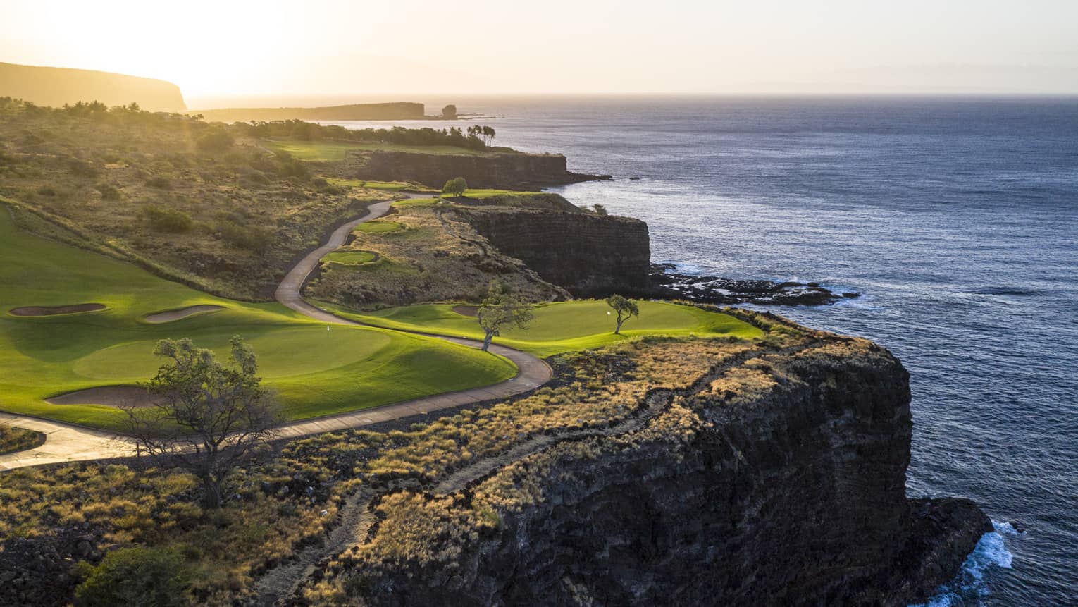 Soft sunlight filters over a pristine golf course with a winding road set on the edge of a cliff overlooking the calm ocean.