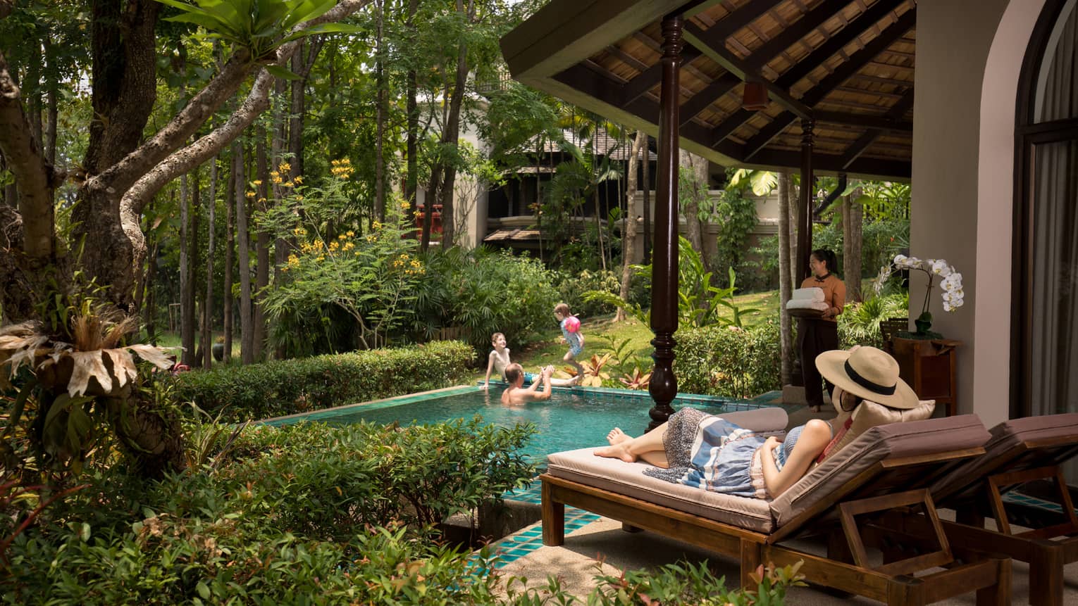 Person relaxing on a sun lounger beside a private pool while others play in the water and a staff member carries fresh towels nearby amidst lush greenery