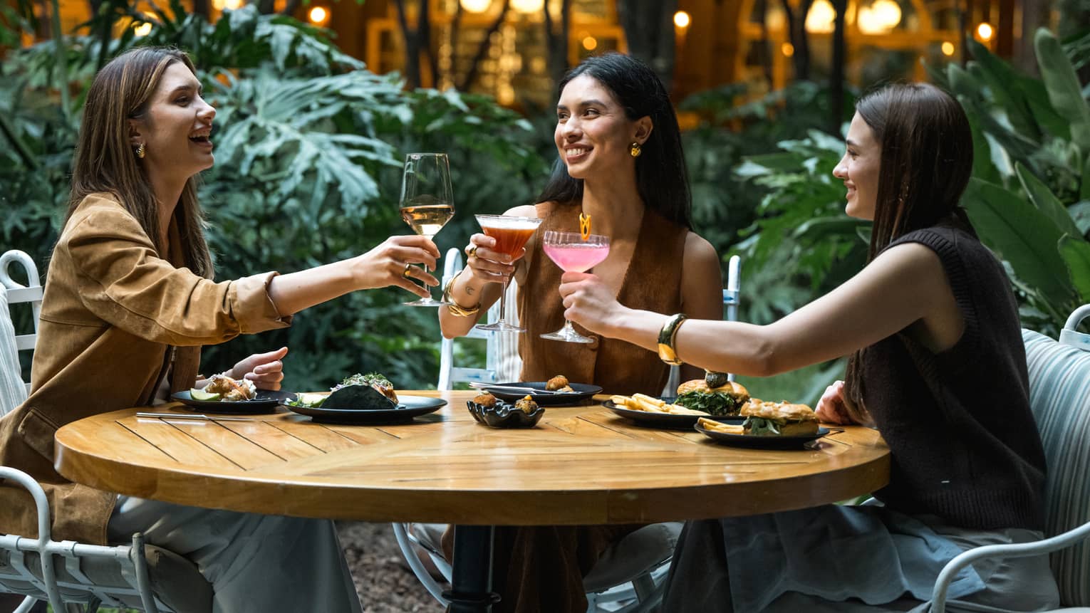 Three guests sit around a round wood table in an outdoor dining space and raise their glasses in a toast.