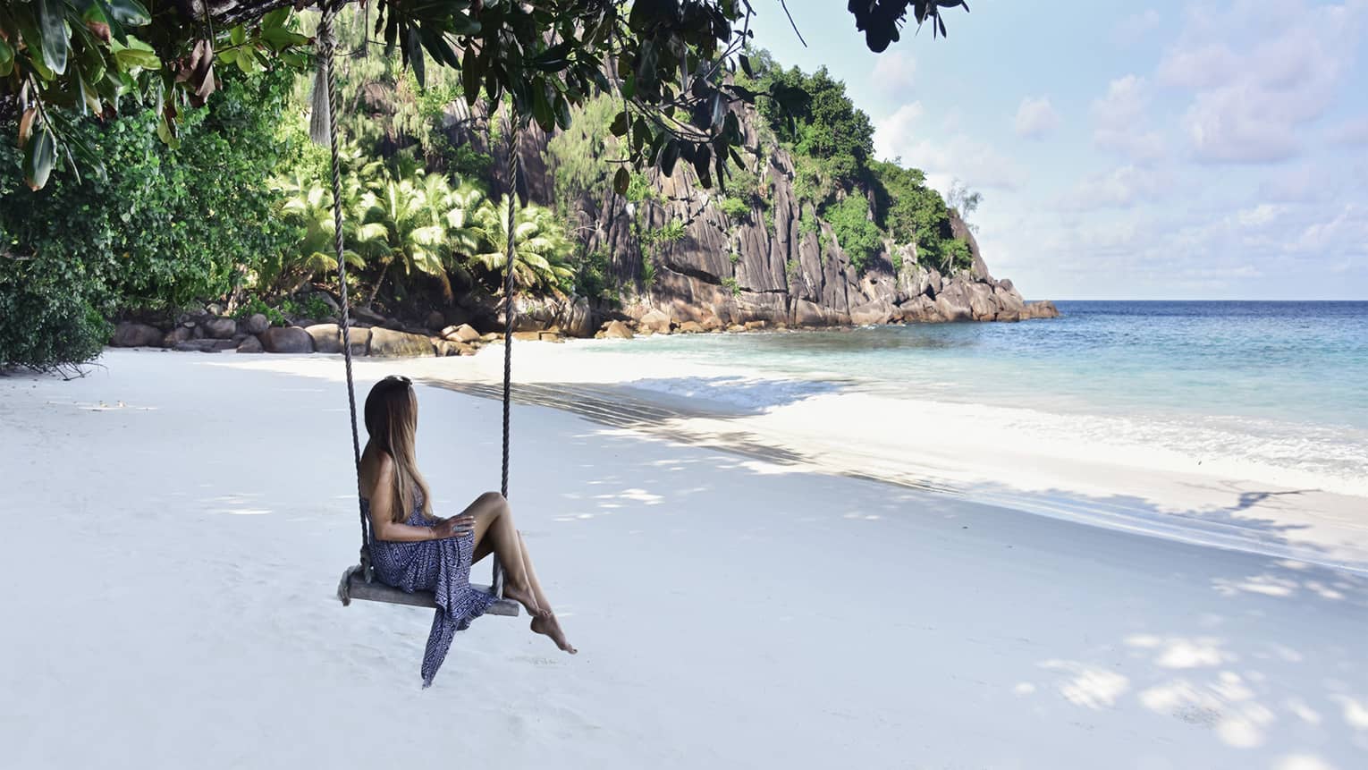 A woman sits on a tree swing on the beach with a view of a cliffside and the ocean