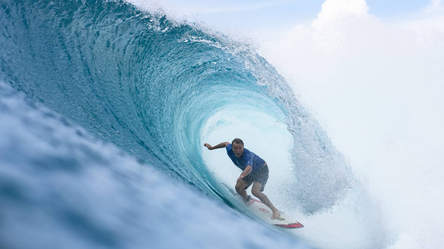 Close-up of a rippling, crystal-clear wave curling over an open-mouthed surfer crouched on a board under a cloudy sky.