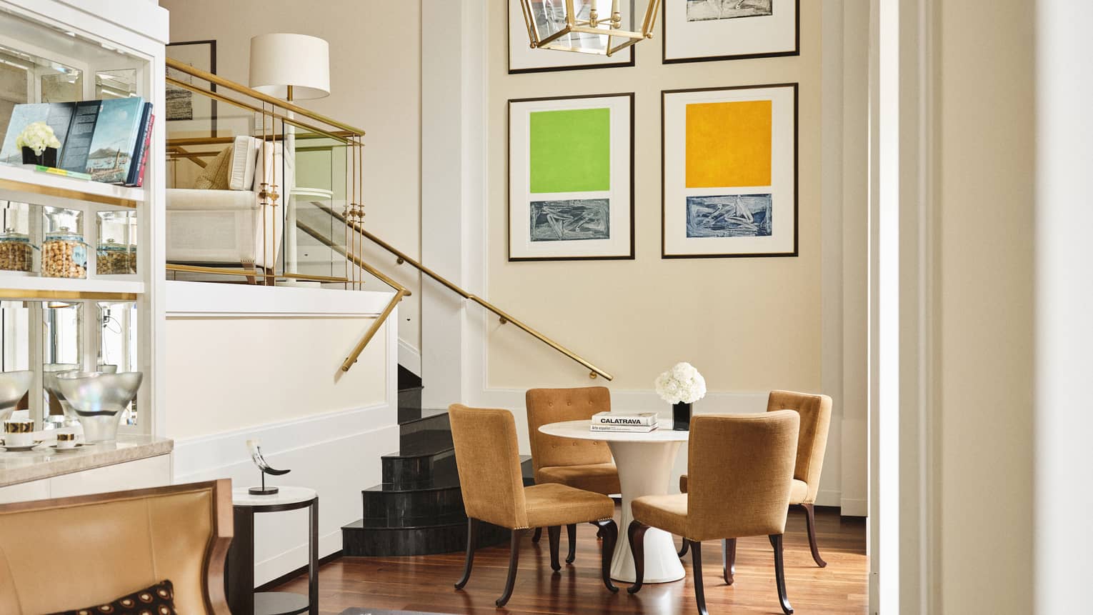 Loft suite dining nook with mustard-coloured chairs around a white round table near a staircase with gold railing and vibrant abstract artwork on the wall