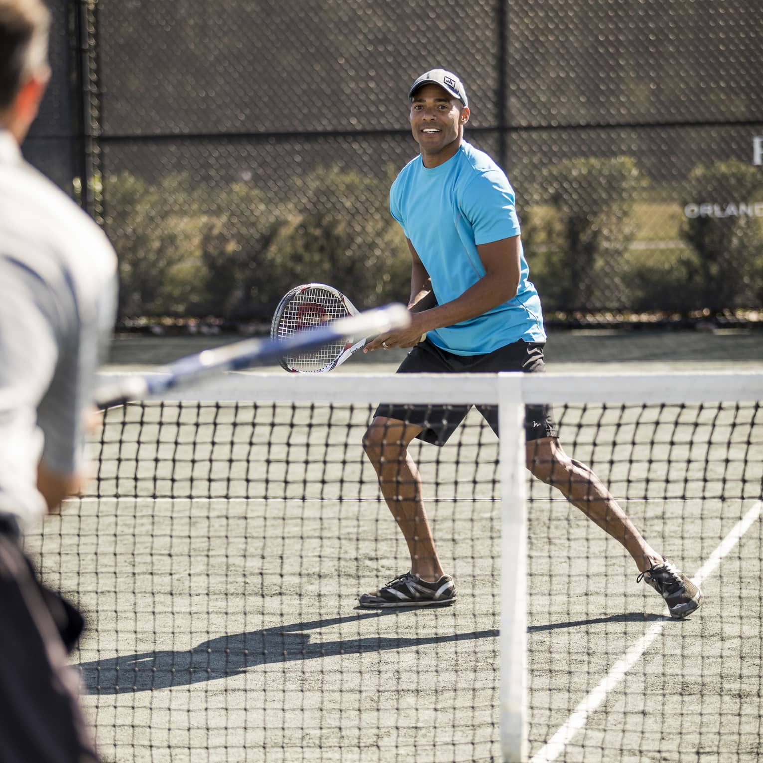 Man wearing blue T-shirt and hat holds racket as tennis ball flies over net