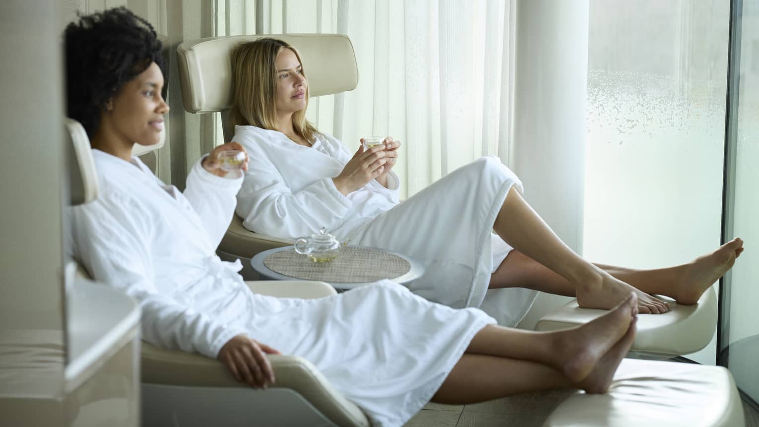 Two spa guests in white robes sitting on lounge chairs drinking champagne