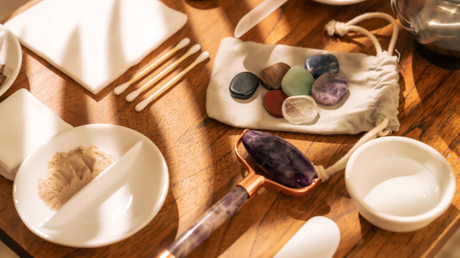 Face roller, gemstones, cotton swabs and a variety of small white bowls and other instruments sit on a sun-lit wooden table
