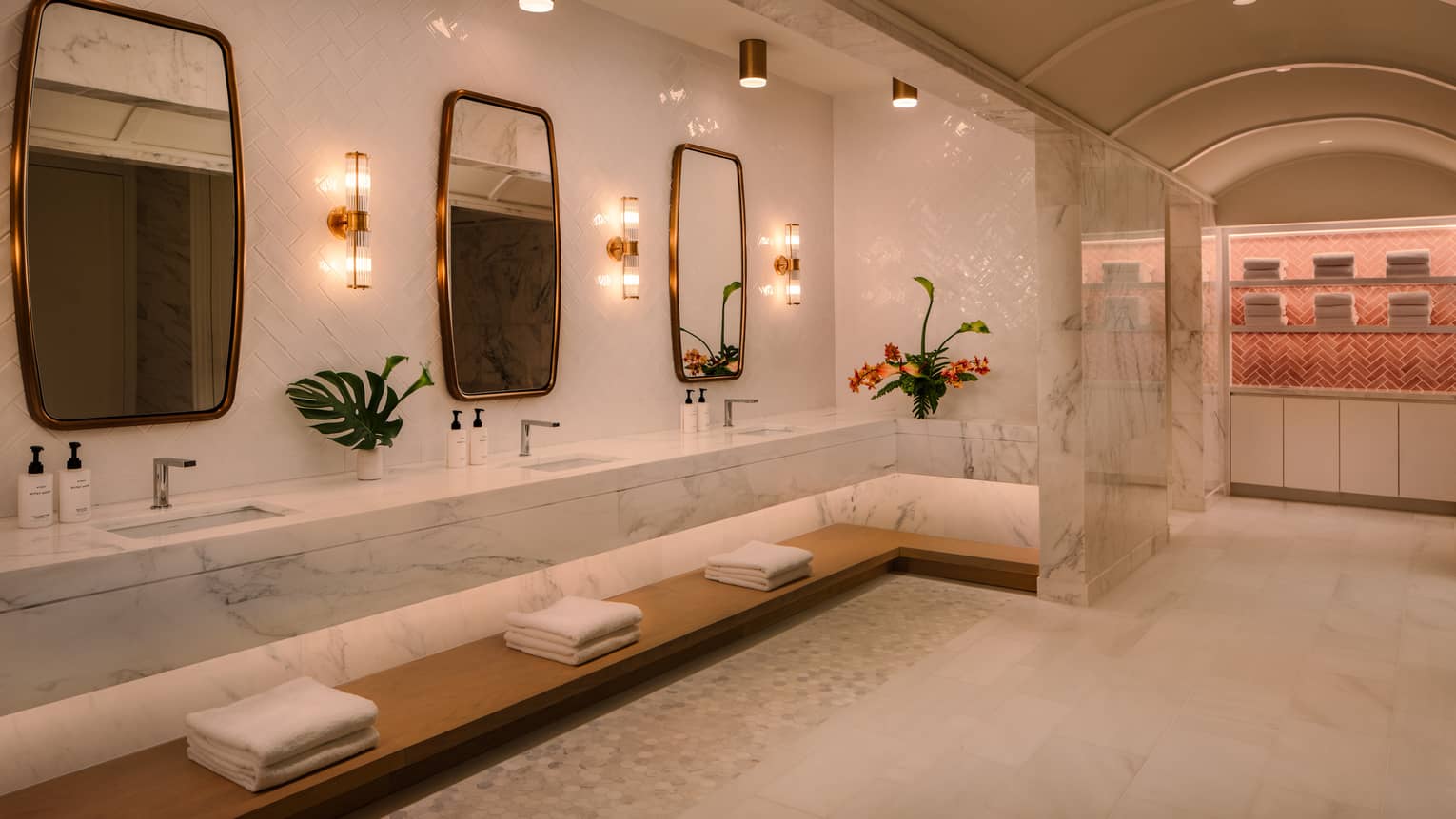 White marble spa bathroom with a long white marble vanity featuring multiple sinks and gold-rimmed mirrors