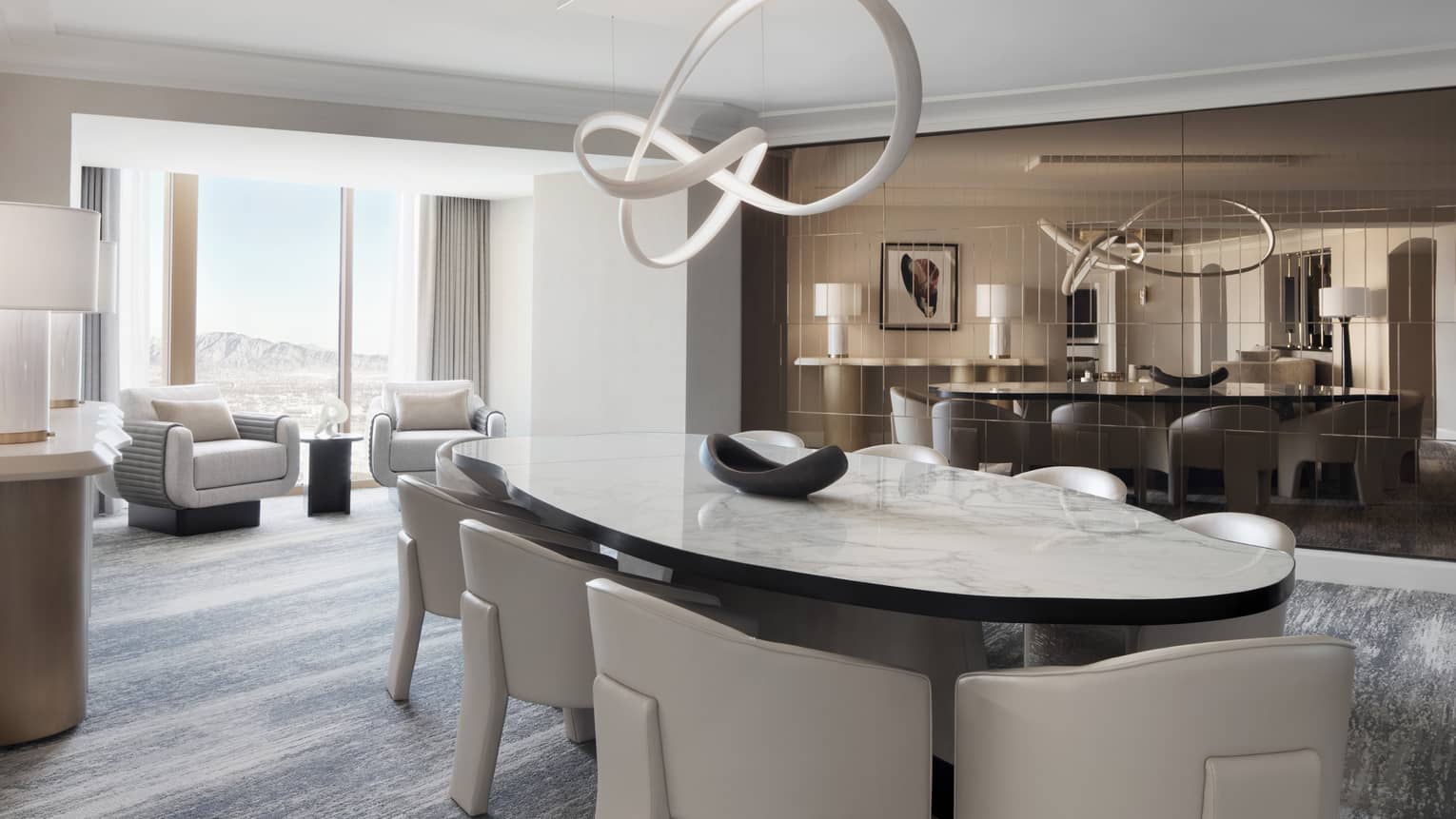 A modern dining room with white upholstered dining chairs and rounded marble table, an abstract chandelier and seating area behind by a large window overlooking a mountain view