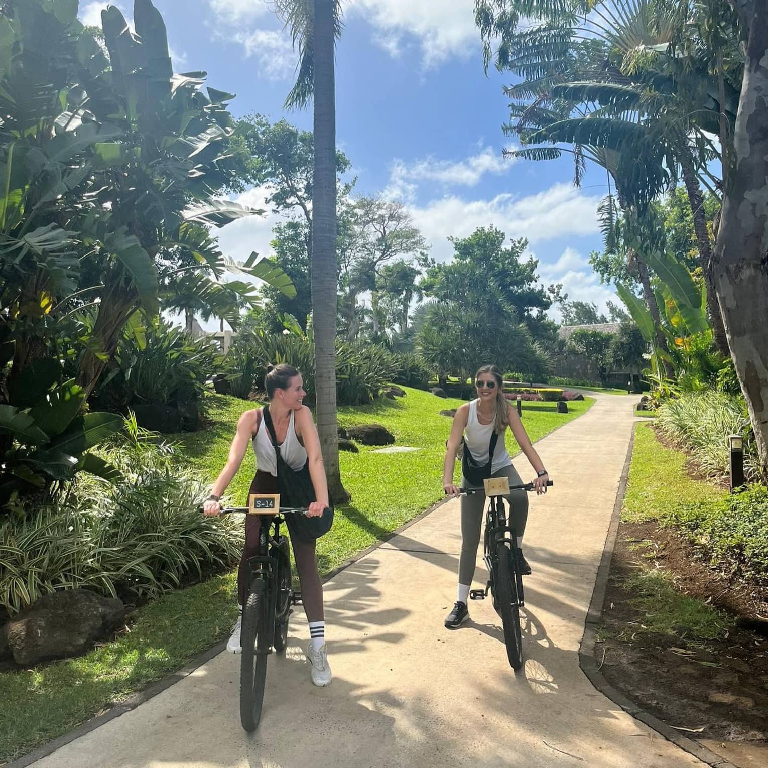 Two guests dressed in athletic attire ride bicycles on a tree-lined path