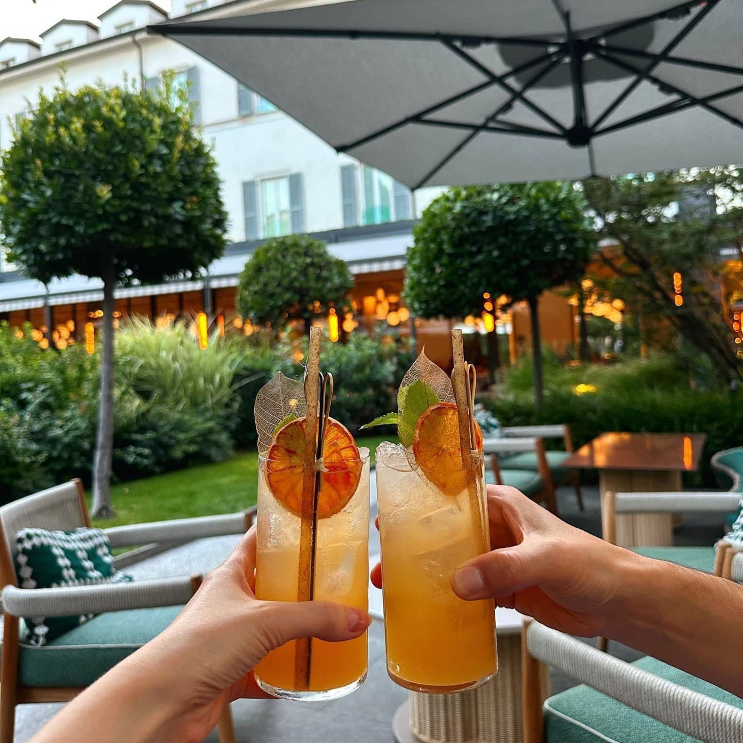 Two hands holding orange cocktails garnished with dried orange slices beneath an umbrella on an outdoor patio at the Four Seasons Hotel Milan