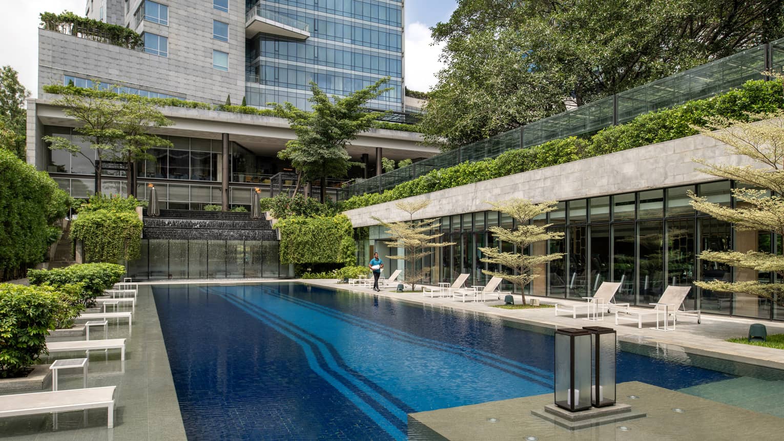 Outdoor lap pool flanked by lounge chairs in the shade of a grey tiled building with a wall of windows