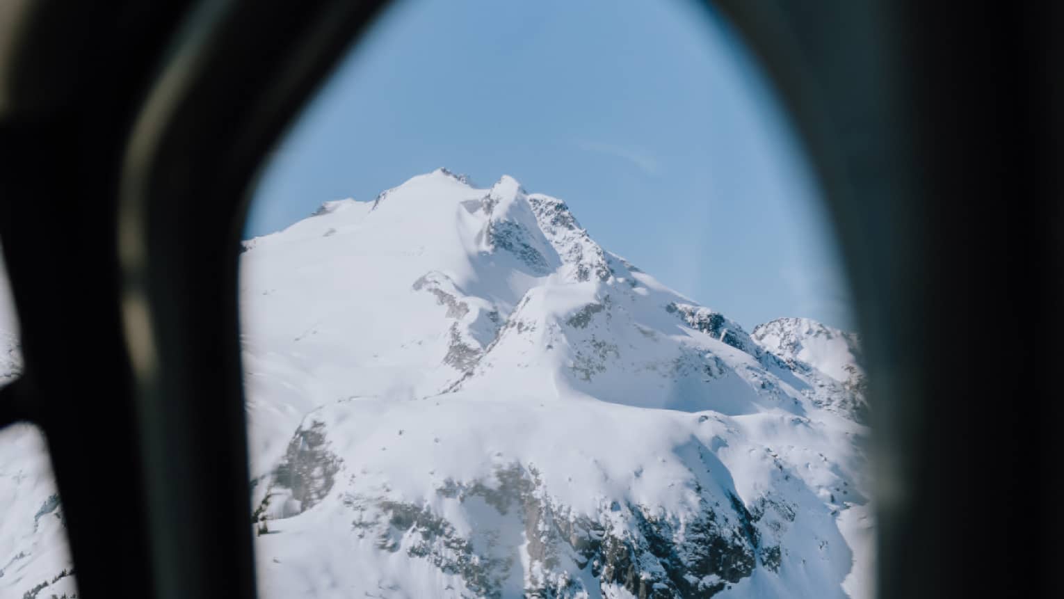An aerial view through an airplane window of a mountain covered in snow with sporadic clumps of trees around the base.