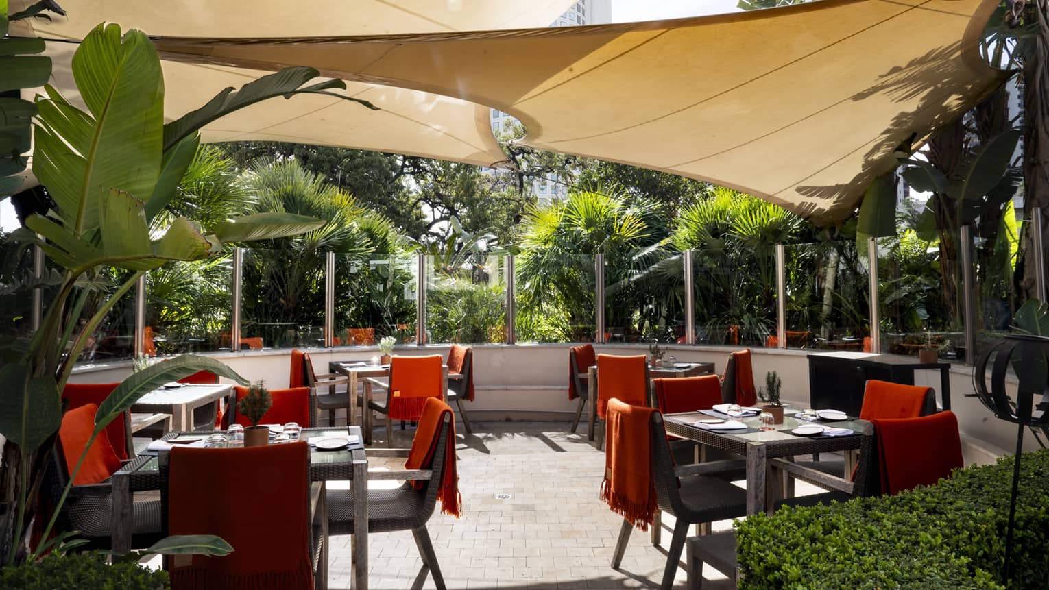 Outdoor dining patio with black-and-red chairs and square tables with three beige sun shades hanging overhead and lush greenery surrouding the patio