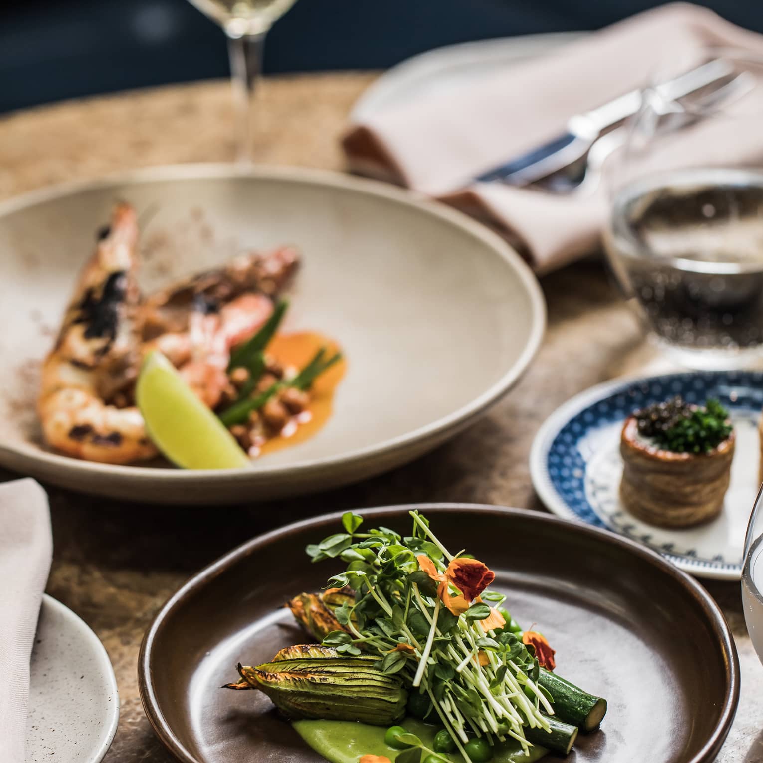 A dish of grilled prawns, a glass of wine, a plate of green peas and roasted zucchini flowers garnished with charred truffle.