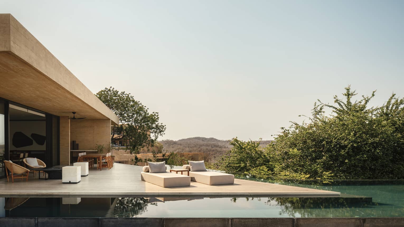 A modern luxury villa with an expansive deck featuring lounge chairs, an outdoor dining area and an infinity pool, offering panoramic views of rolling hills and greenery