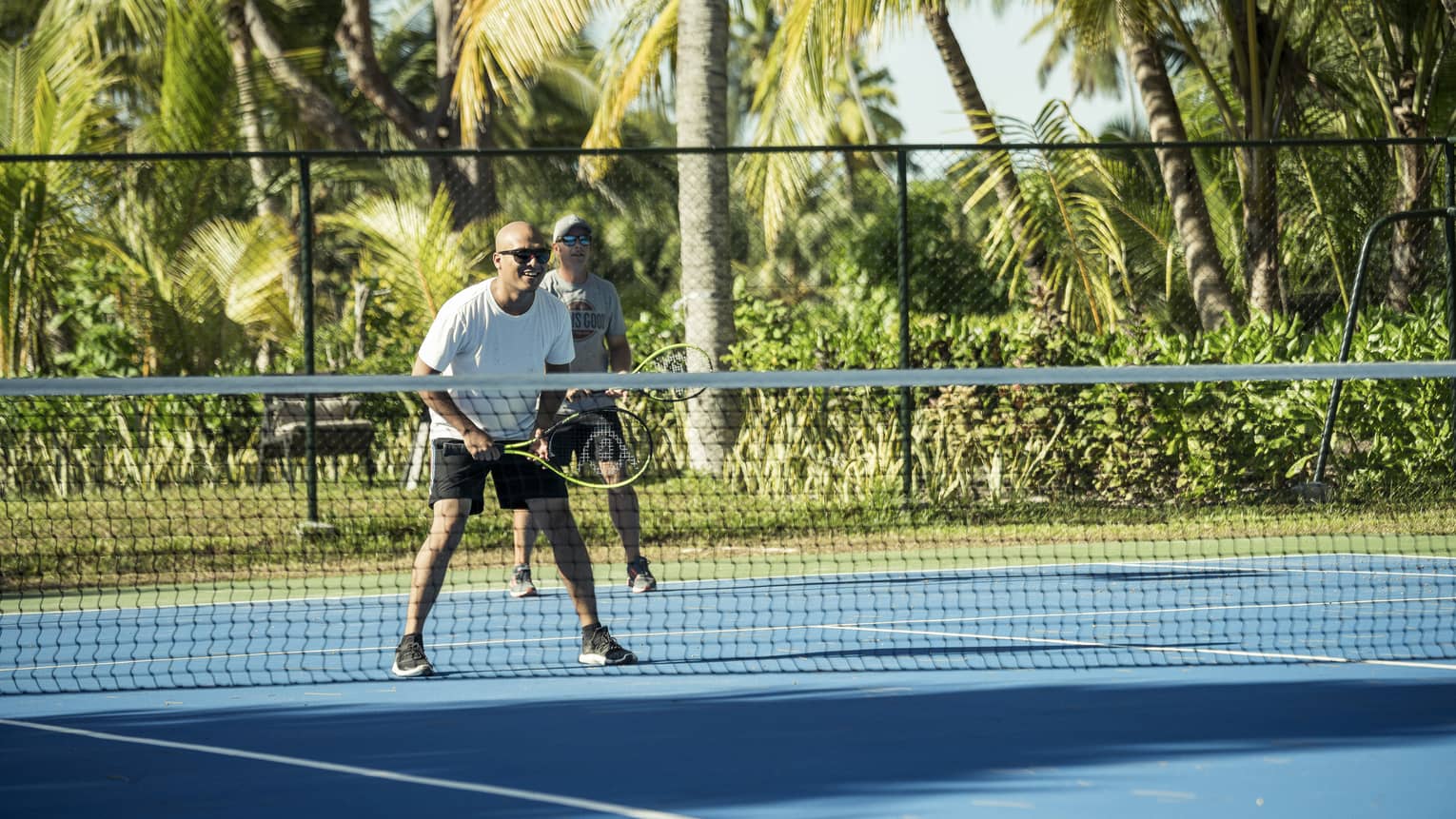 Two tennis players stand on the same side of the court with their racquets aloft, lush greenery and palm trees behind them.