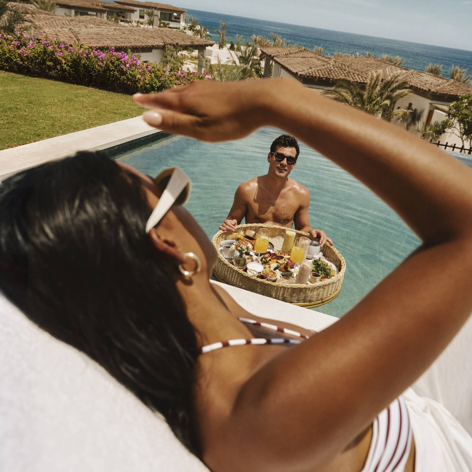 A guest stands in a pool with a floating basket of breakfast food and drinks and looks up at another guest sitting poolside.