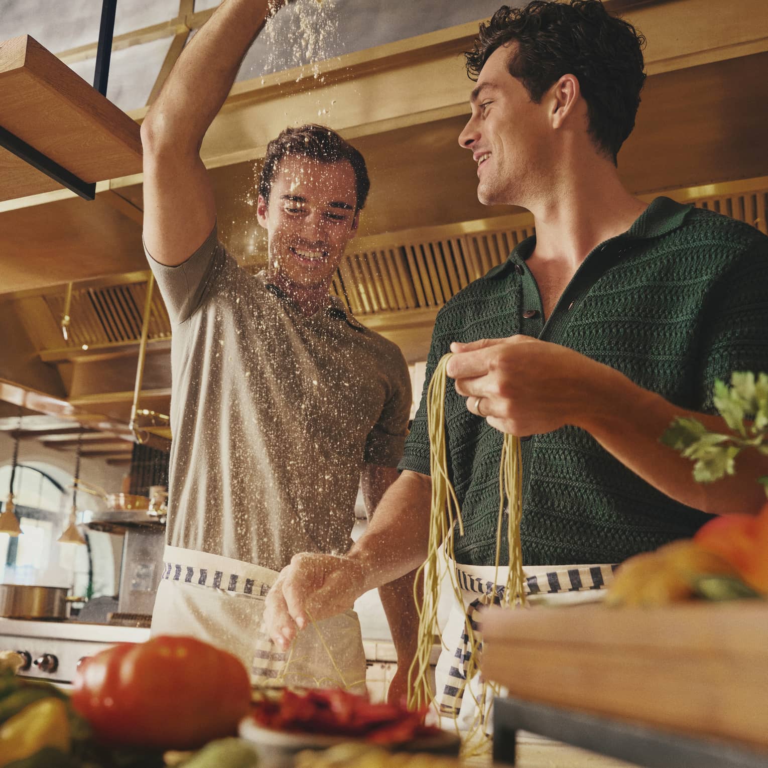 Two smiling guests in aprons prepare fresh pasta noodles on a counter laden with vibrant fresh produce in a bright kitchen.