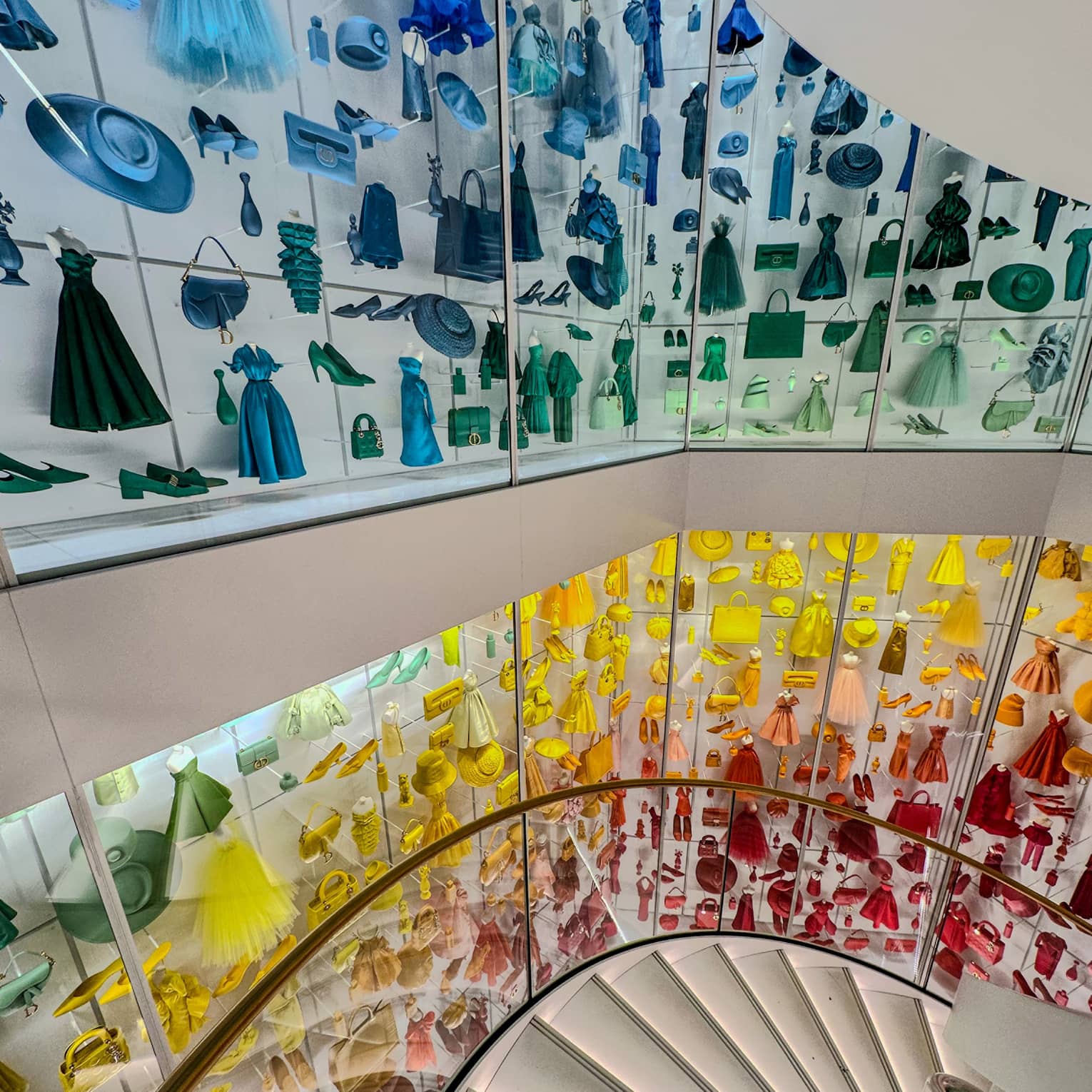 View down a circular staircase lined with glass display cases showcasing various Dior pieces in cascading rainbow colours.