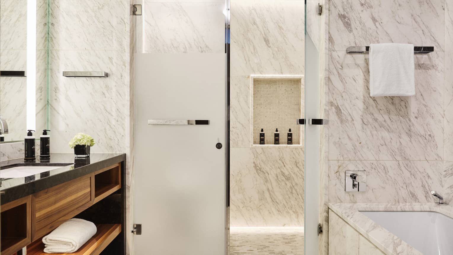 Luxury marble bathroom with walk-in shower, bathtub, vanity with black countertop and wood shelving, towel rack and toiletries displayed on a recessed shelf