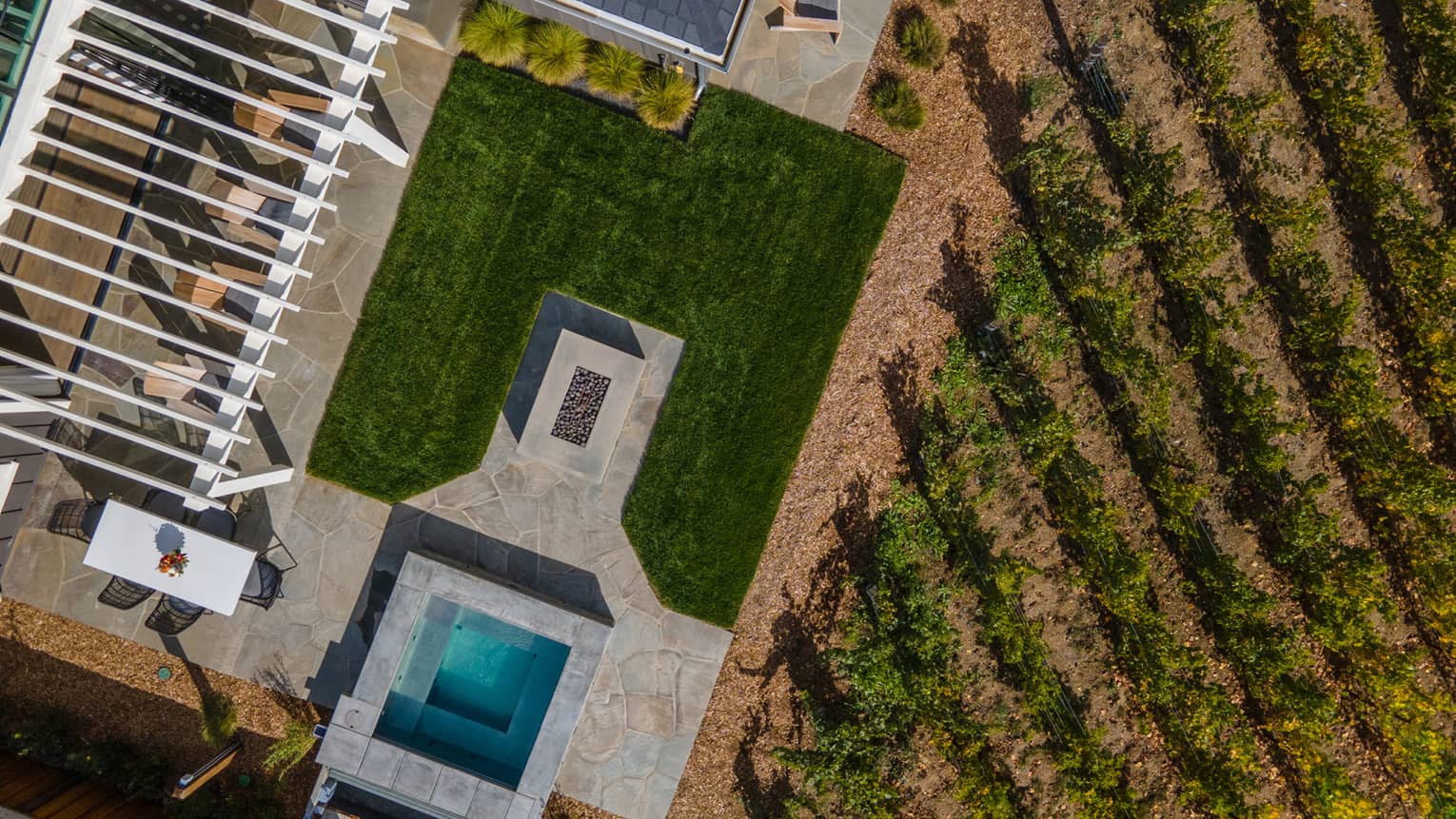 Aerial view of a luxury resort villa patio with a pergola, dining table, fire pit, hot tub, green lawn and adjacent vineyard rows