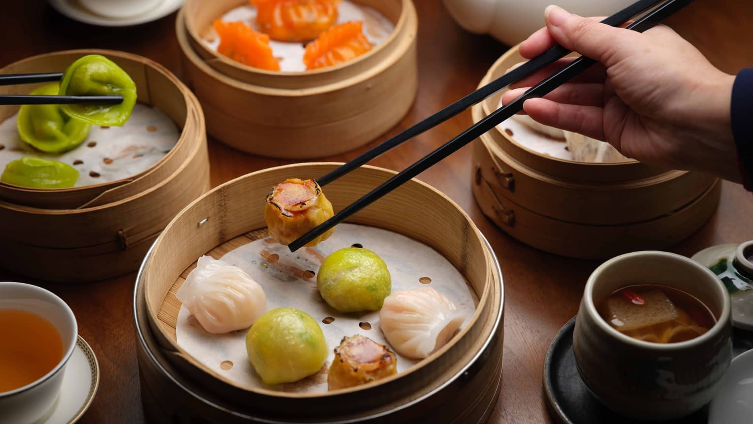 Beside cups of tea, a hand uses chopsticks to pick up one of the varied pieces of dim sum picked from nearby bamboo steamers.