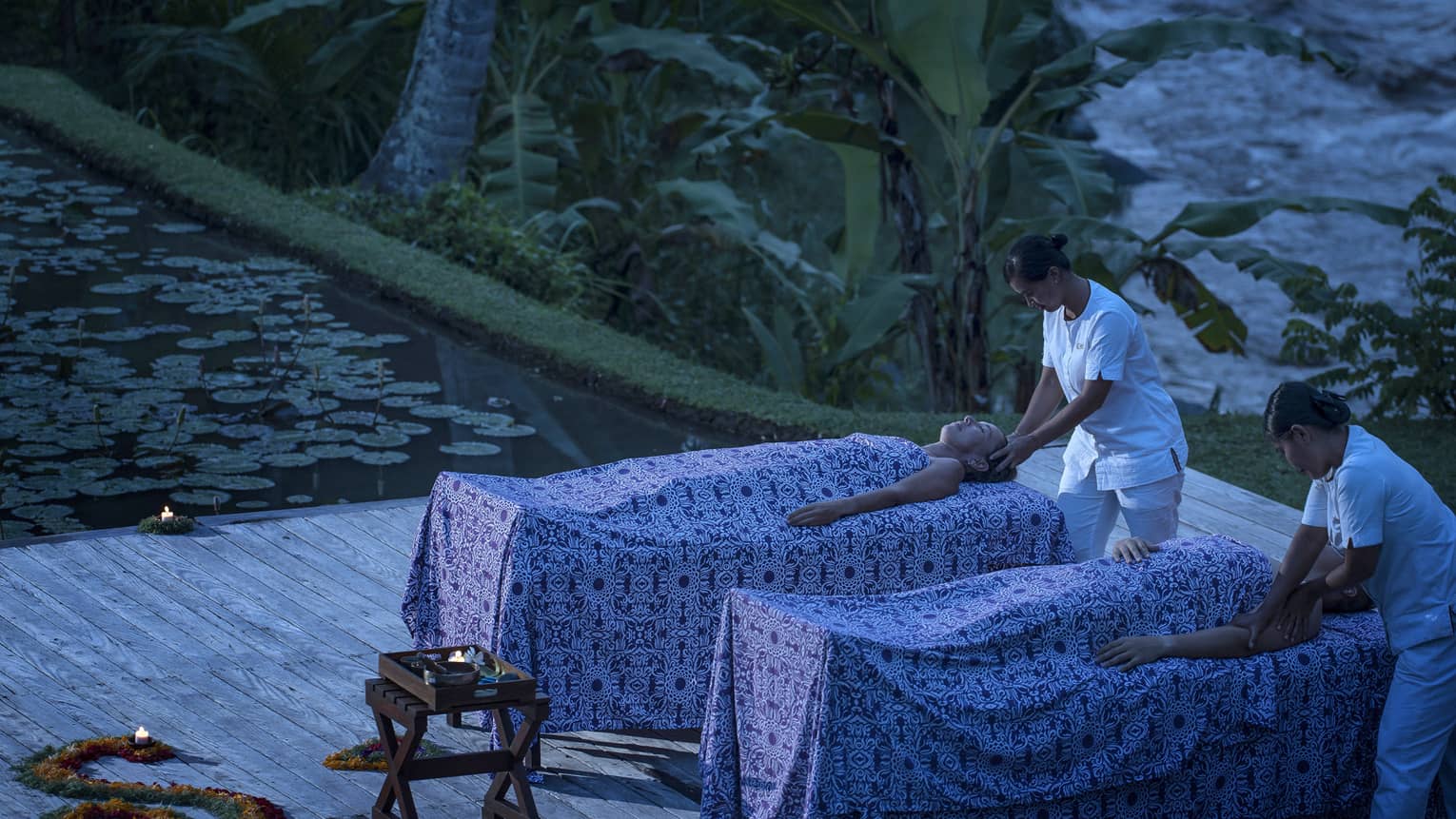 Two guests receive massages by candlelight beside an outdoor pool of lotus and a backdrop of tropical greenery.