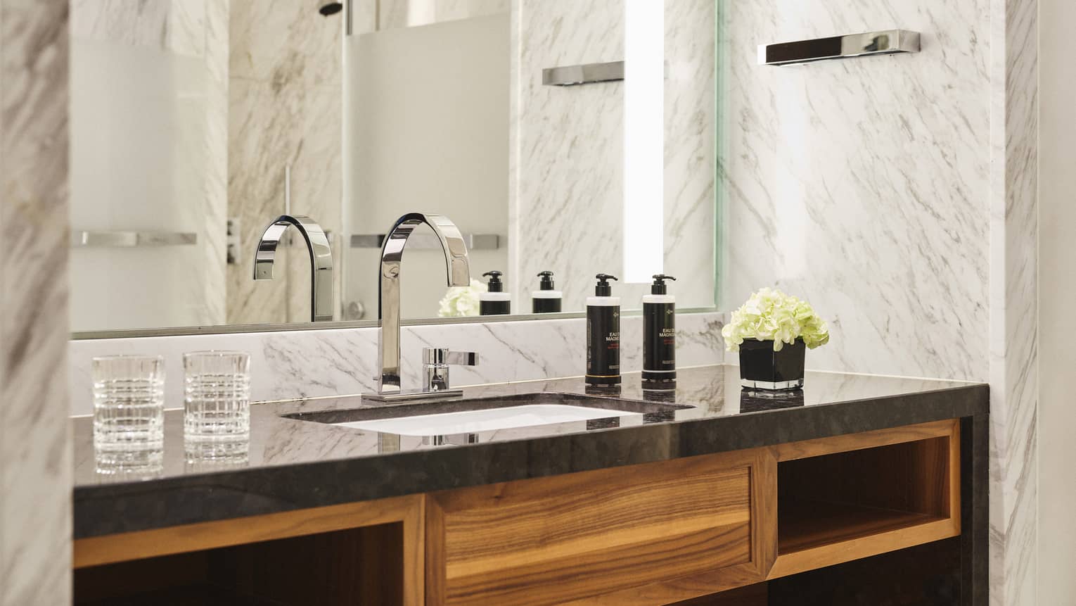 Hotel bathroom vanity with black countertop, chrome faucet, wooden shelving, two glasses, toiletries, a small bouquet of white flowers, mirror and marble walls