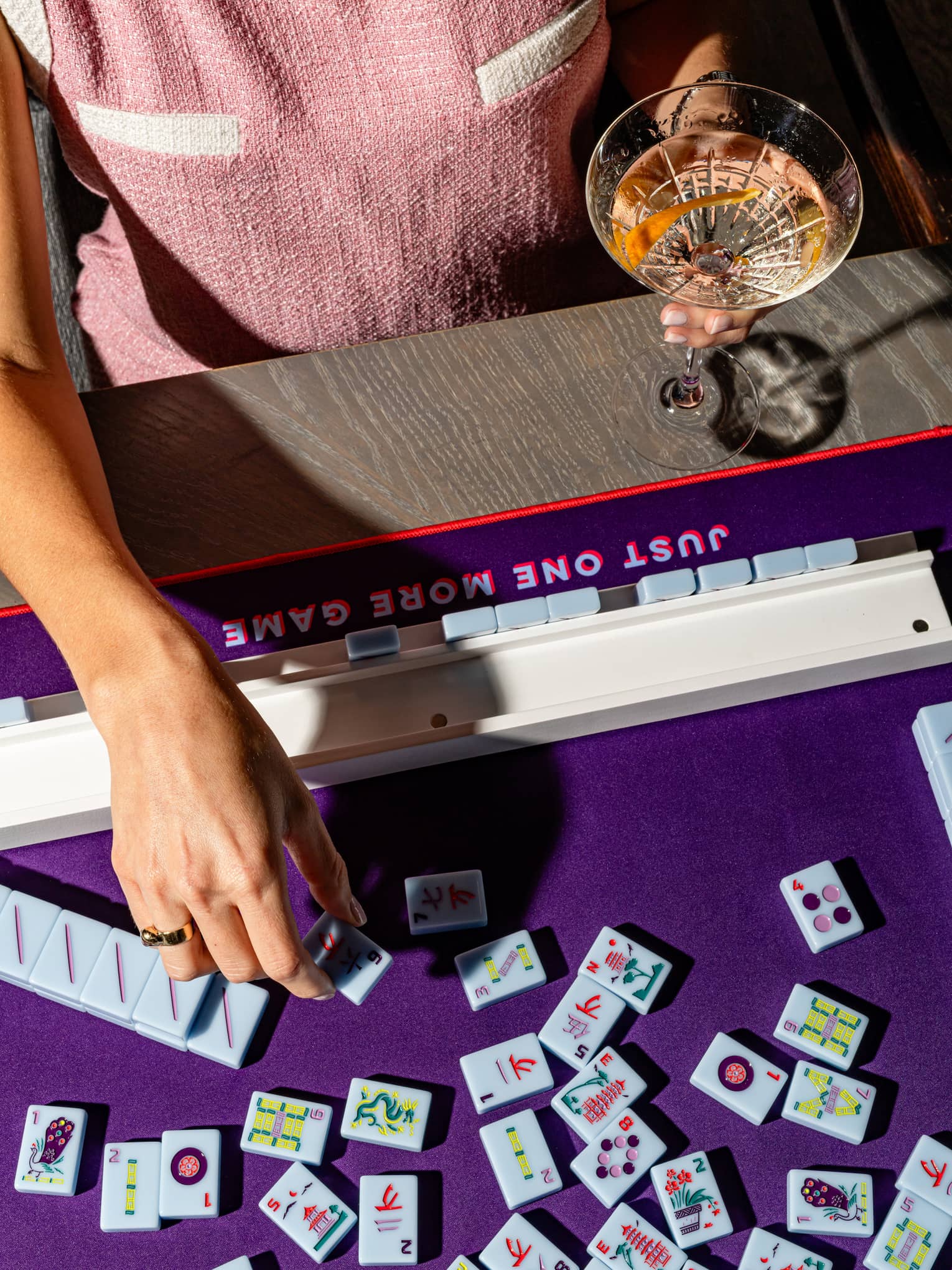 Person wearing a pink-and-white dress and holding a cocktail in one hand plays mahjong on a violet table