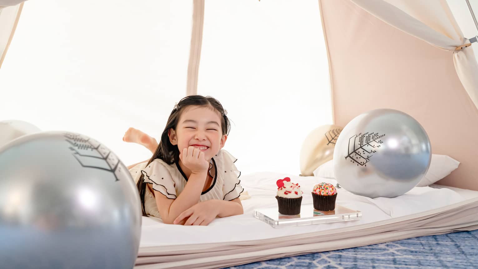 Smiling young girl in in-room kids? tent with gold and silver balloons and cupcakes