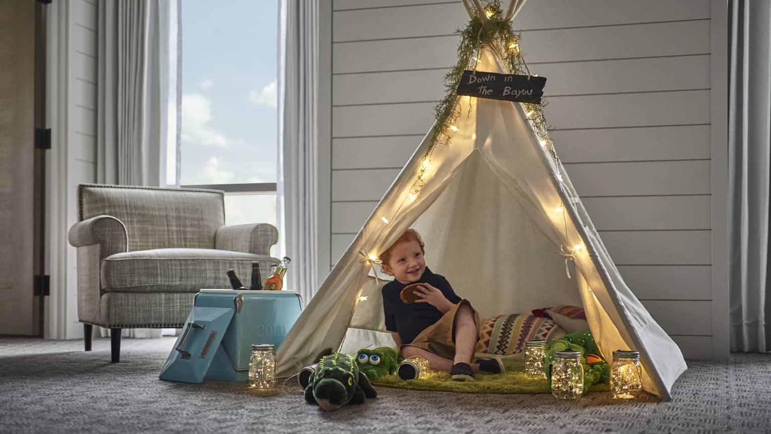 In a small tepee, a smiling child holds a cookie and is surrounded by pillows, stuffed animals and jars of twinkling lights.