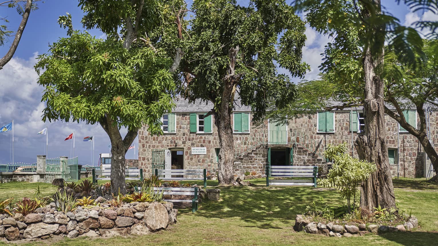 A two-storey stone building with green doors and shutters, seen across a lawn and behind trees, flags blowing behind it.