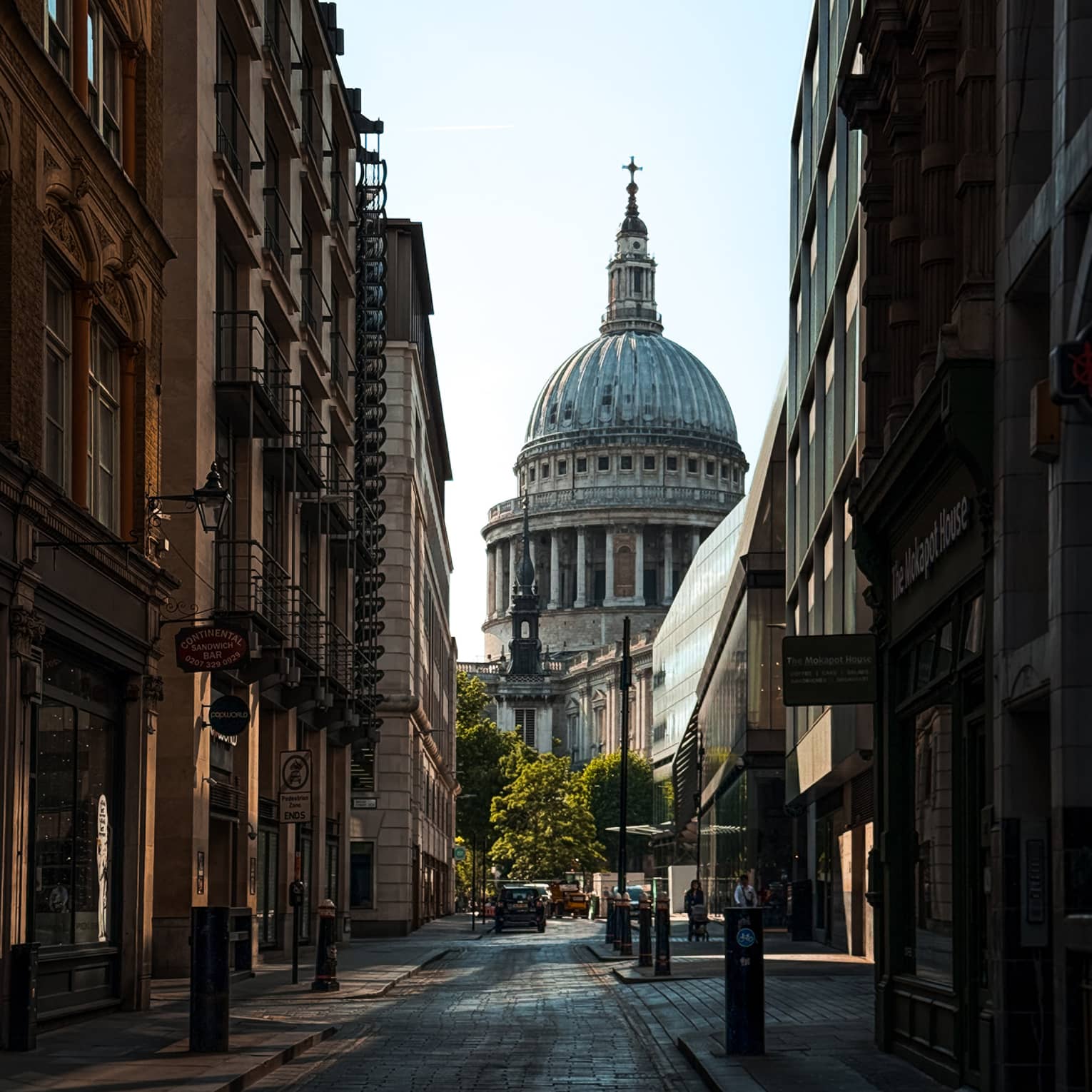 A city street in shadow, lined with buildings. The dome and spire of a cathedral rise in the background, beyond the end of the street.