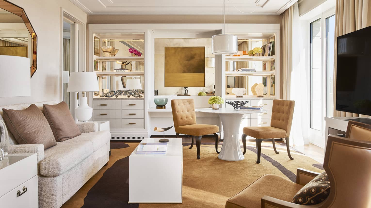 Luxury hotel living room with plush seating, tan chairs, round and rectangular tables, TV, decorative shelves and large windows with beige curtains