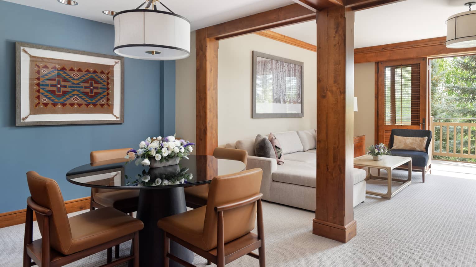 Dining area with a round glass table, leather chairs, and a blue accent wall displaying a framed textile art piece, opening to a living room with a sectional sofa