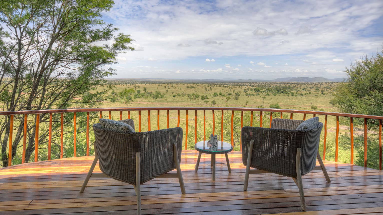 Villa wooden balcony with two wicker chairs separated by small round table and overlooking expansive plain through vertical wooden railing