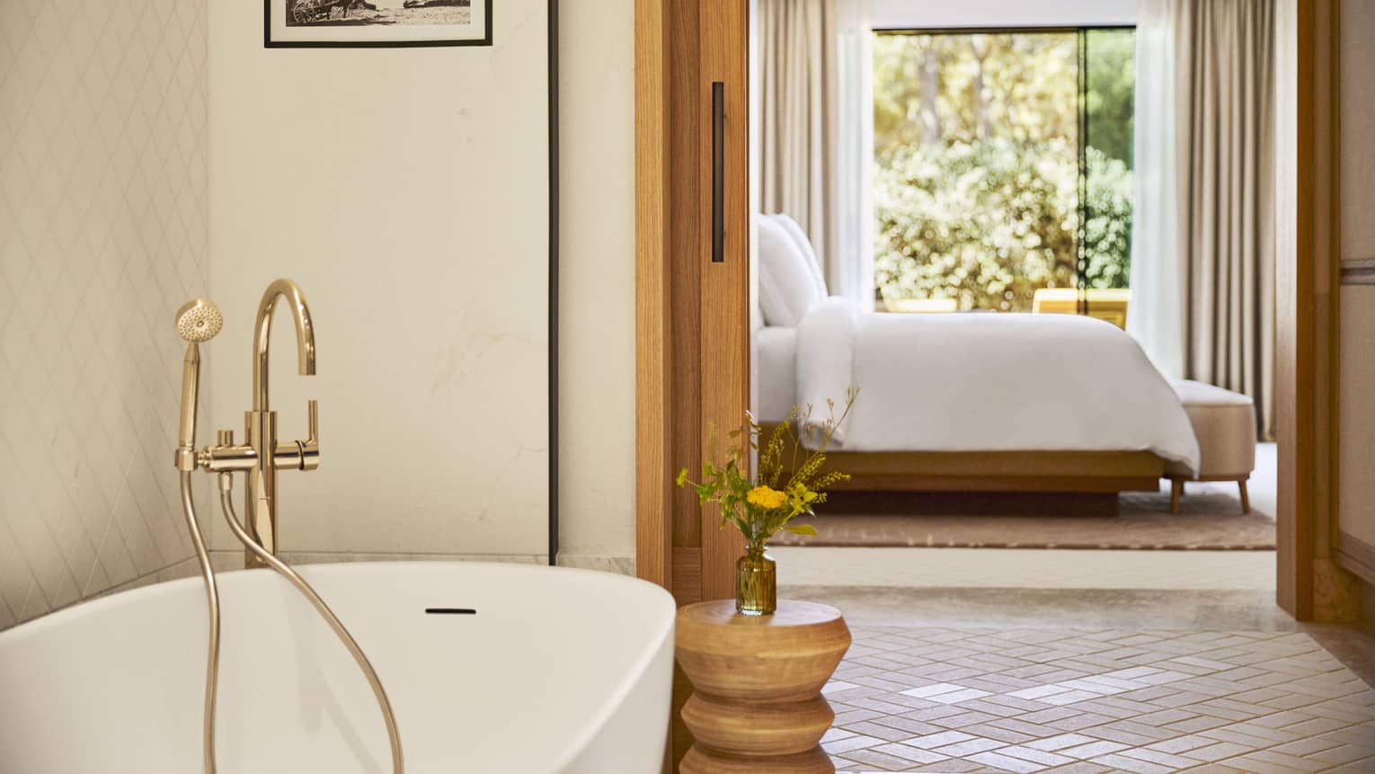 View from a bathroom with brass fixtures on a luxe soaker tub toward a sunlit bed made up in fresh white linens.
