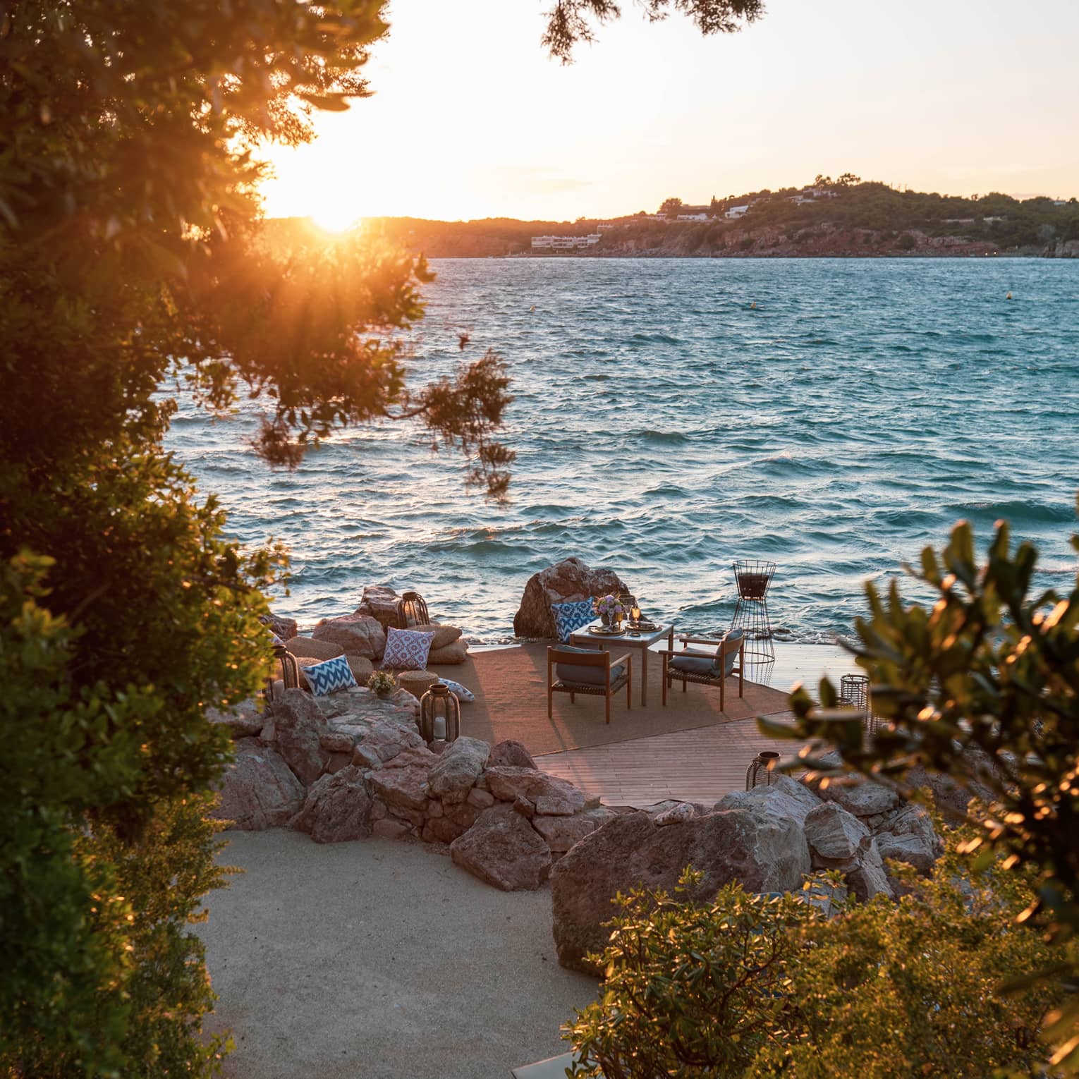 From tree-lined stairs, a furnished patio, including table set for two beside rippling water and sun setting over a distant isle