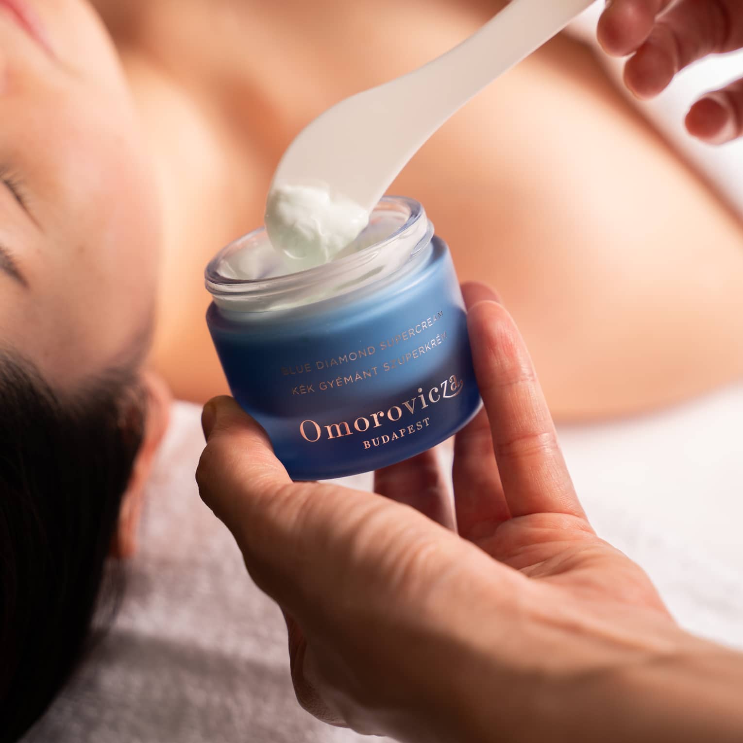 Hand dips spatula into jar of Omorovicza face cream in preparation for woman’s facial