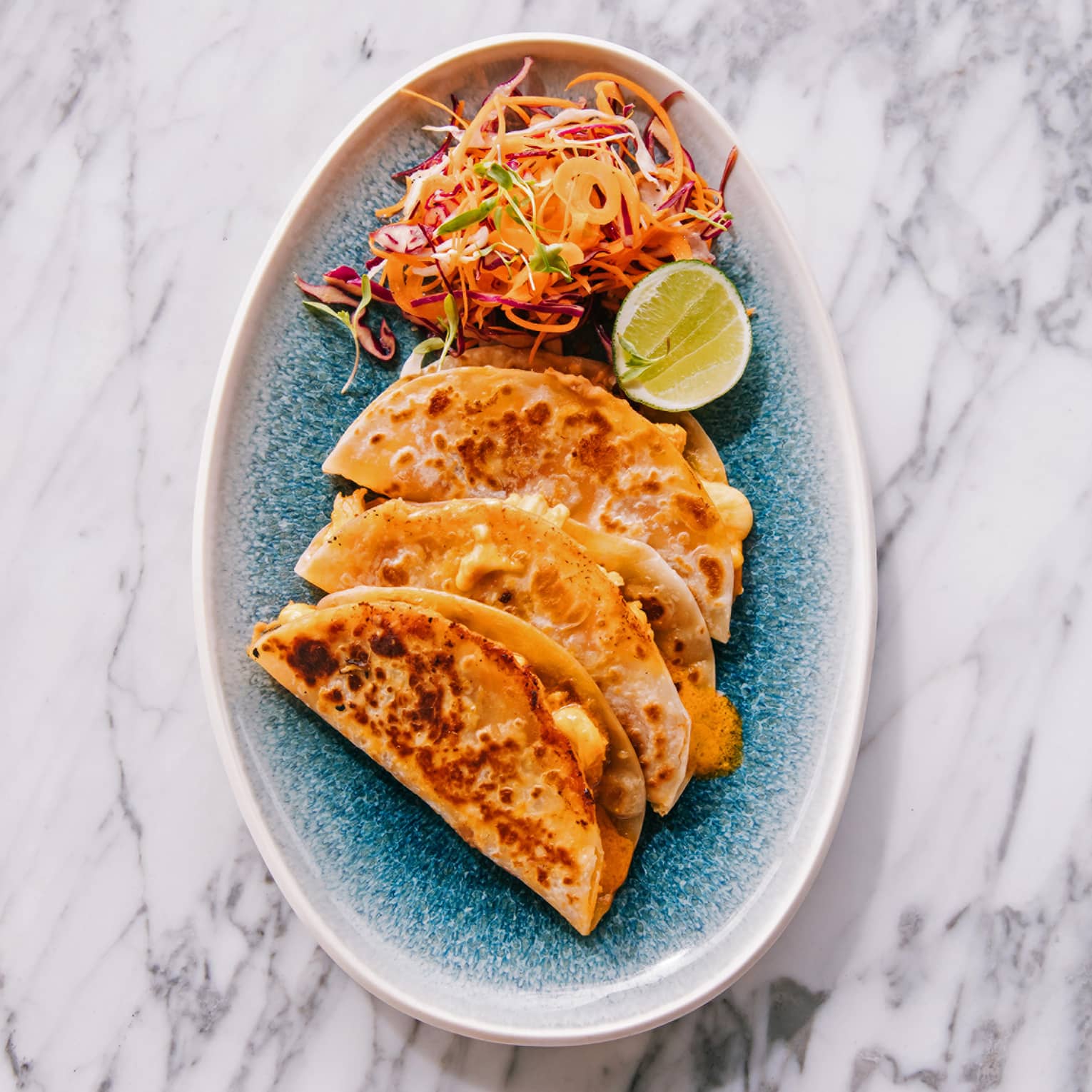 Three tacos and a brightly colored salad with a slice of lime on a blue plate create a visually appealing and appetizing dish.