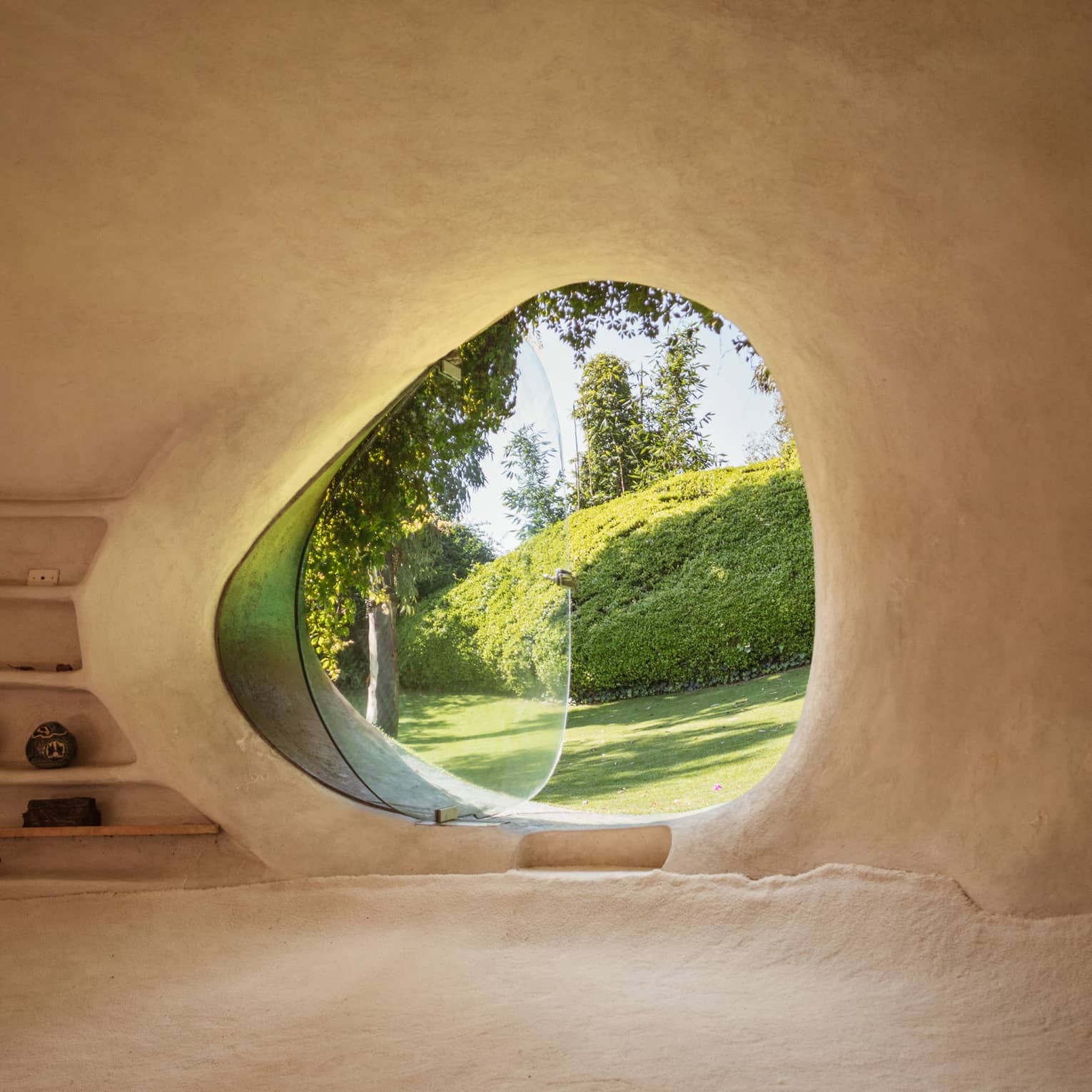 Smooth, adobe wall frames a curved, asymmetrical window, glass pivoted out, a verdant yard with large pruned hedge in view.