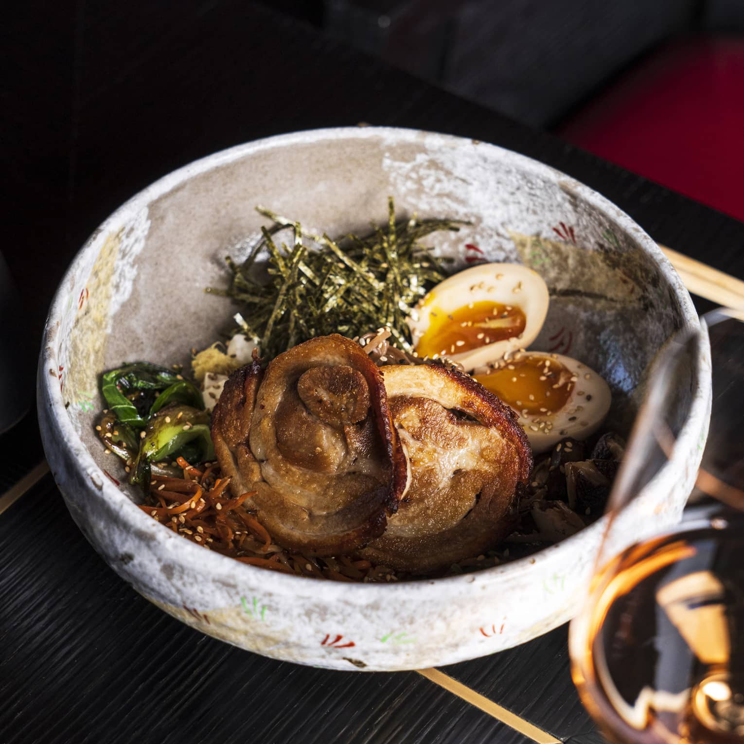 A stone bowl full of roasted meat, softboiled eggs, seaweed strips and vegetables sprinkled with sesame seeds.