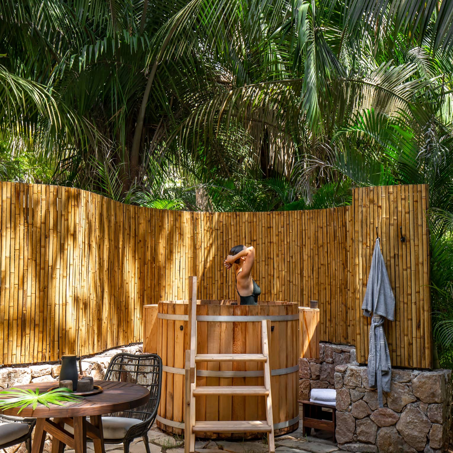 Woman emerges from soak in natural wooden outdoor bath, bamboo fence and palm trees surrounding
