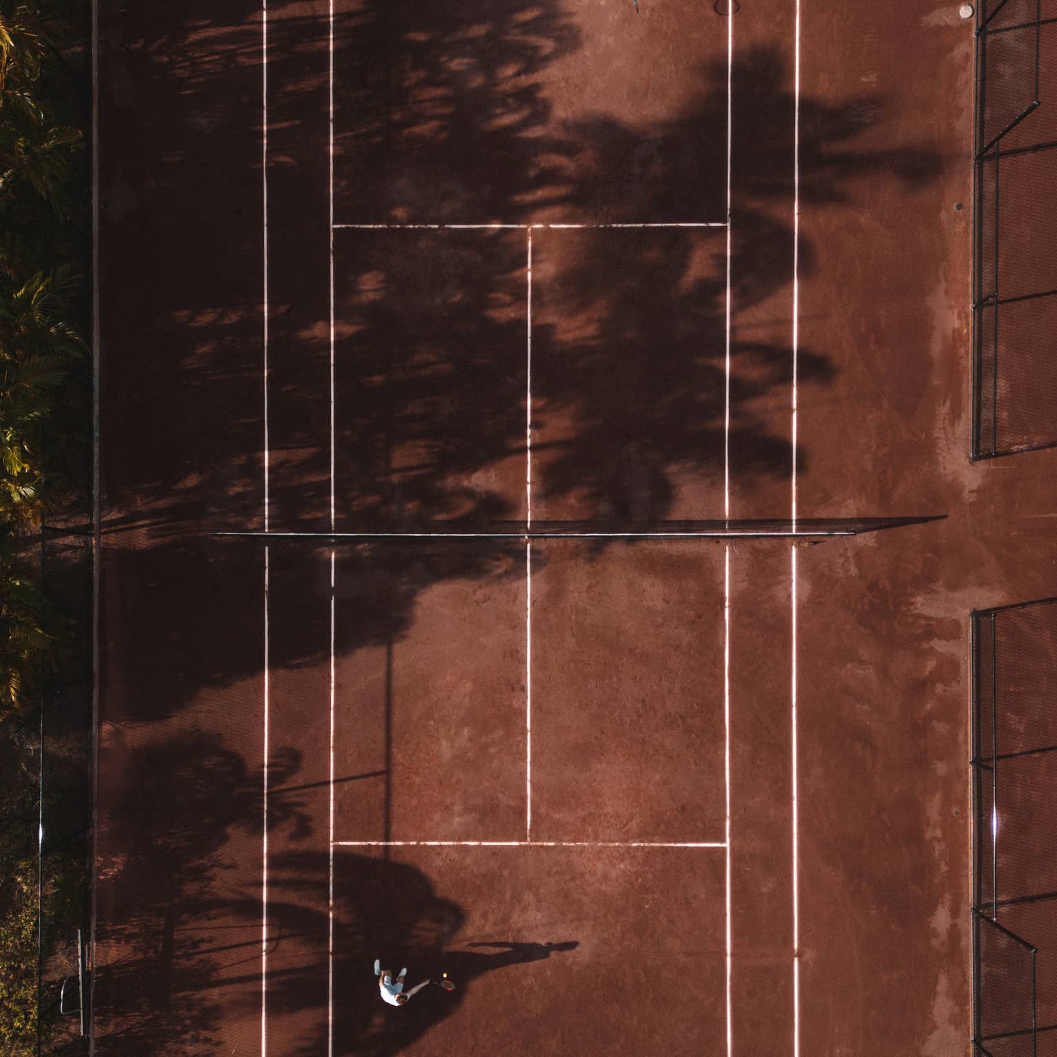 An aerial view of a red clay tennis court partially shaded by trees