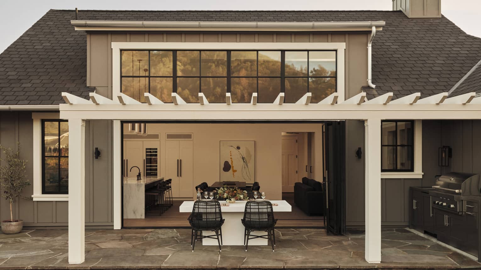 Outdoor dining area of a residential villa with pergola, white table and black wicker chairs, opening to a kitchen and living room through large folding doors